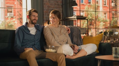 Portrait of Young Couple Spending Time at Home, Sitting on a Sofa, Man Embracing His Girlfriend While Watching Exciting TV Show in Apartment. Man and Woman Streaming Movie and Get Surprised.