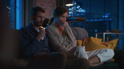 Portrait of Beautiful Couple Spending Time at Home, Sitting on a Couch, Watching Scary TV Show in Their Stylish Loft Apartment. Man Puts Hand on Female Knee, While Streaming Thriller Movie.