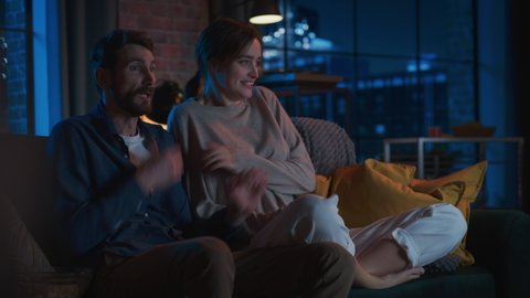Portrait of Beautiful Couple Spending Time at Home, Sitting on a Couch, Watching Scary TV Show in Their Stylish Loft Apartment. Man and Woman Streaming Horror Movie and Get Scared and Surprised.