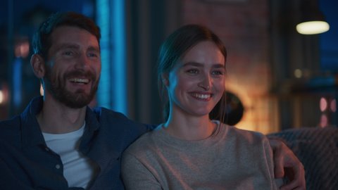 Portrait of Beautiful Couple Spending Time at Home, Sitting on a Couch, Hugging and Watching Exciting TV Show in Their Stylish Loft Apartment. Man and Woman Streaming Comedy Movie and Have Good Time.