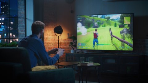 Young Man Spending Time at Home, Sitting on a Couch in Loft Apartment and Playing Arcade Shooter Video Game on Console. Male Using Controller to Play MMO Battle Royale Style Game with Virtual Friends.