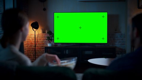 Young Couple Spending Time at Home, Sitting on a Couch and Watching TV with Green Screen Mock Up Display in Their Stylish Loft Apartment. Man and Woman Stream Movie or Show. Shot From Back at Night.