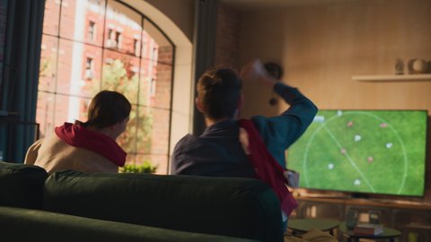 Couple of Soccer Fans Relax on a Sofa, Watch a Sports Match at Home in Stylish Loft Apartment. Excited Young Man and Woman Cheer for Their Favorite Football Club, Celebrate Player Scoring a Goal.