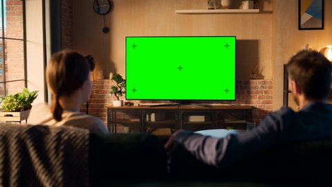 Authentic Couple Spending Time at Home, Sitting on a Couch and Watching TV with Green Screen Mock Up Display in Their Stylish Loft Apartment. Man and Woman Streaming Movie or Show. Shot From Back.