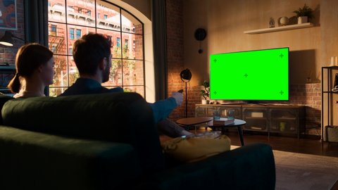 Young Couple Spending Time at Home, Sitting on a Couch and Watching TV with Green Screen Mock Up Display in Their Stylish Loft Apartment. Man is Switching Channels on Remote Control.