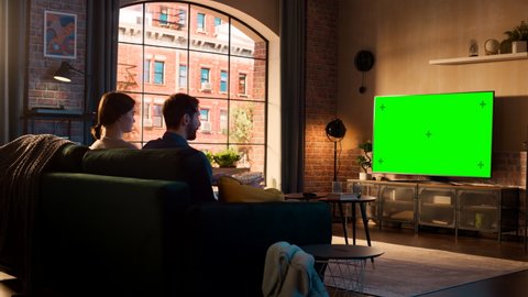 Young Couple Spending Time at Home, Sitting on a Couch and Watching TV with Green Screen Mock Up Display in Their Stylish Loft Apartment. Man and Woman Streaming Movie or Show and Eating Popcorn.