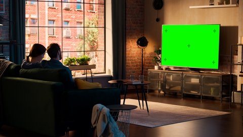 Beautiful Couple Spending Time at Home, Sitting on a Couch and Watching TV with Green Screen Mock Up Display in Their Stylish Loft Apartment. Man and Woman Streaming Movie or Show and Eating Popcorn.