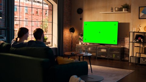 Authentic Couple Spending Time at Home, Sitting on a Couch and Watching TV with Green Screen Mock Up Display in Their Stylish Loft Apartment. Man and Woman Streaming Movie or Show and Eating Popcorn.