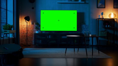 Stylish Loft Apartment Interior with TV Set with Green Screen Mock Up Display Standing on Television Stand. Empty Living Room at Home with Chroma Key Placeholder on Monitor. Zoom Out Evening Shot.