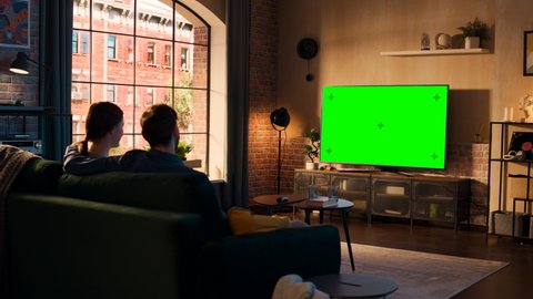 Young Couple Spending Time at Home, Sitting on a Couch and Watching TV with Green Screen Mock Up Display in Their Stylish Loft Apartment. Man and Woman Streaming Movie or Show and Eating Popcorn.