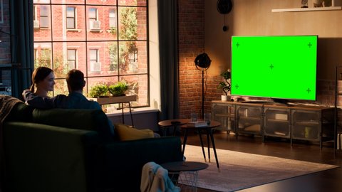 Authentic Couple Spending Time at Home, Sitting on a Couch and Watching TV with Green Screen Mock Up Display in Their Stylish Loft Apartment. Man and Woman Streaming Movie or Show and Eating Popcorn.