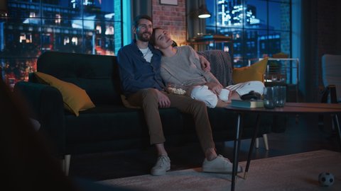 Portrait of Beautiful Couple Spending Time at Home, Sitting on a Couch, Hugging and Watching Exciting TV Show in Their Stylish Loft Apartment. Man and Woman Streaming Comedy Movie and Have Good Time.