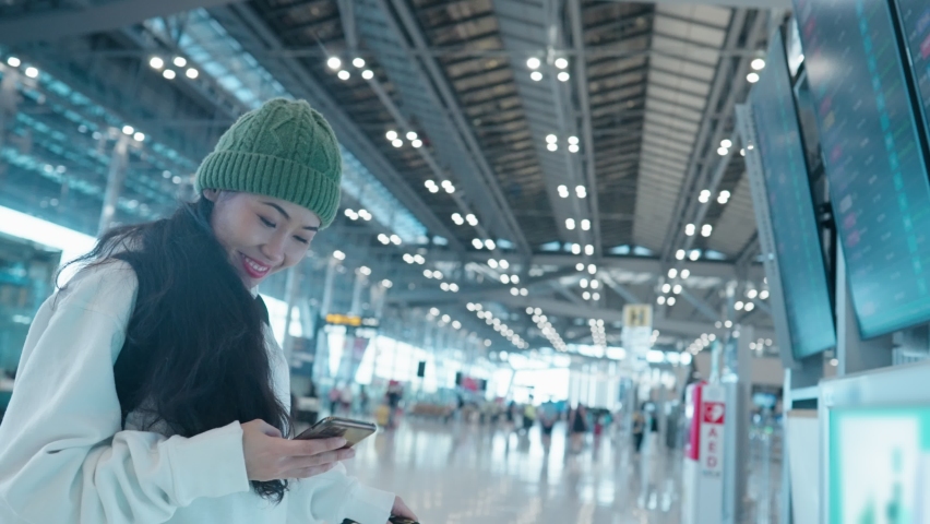 Cheerful asian teenage tourist girl scrolling phone, looking up at the blue airport schedule time table board, traveler in airport terminal, covid-19 pandemic, new normal, flight information, check-in Royalty-Free Stock Footage #1089185575