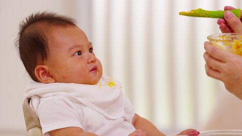 Side view mother feeding her asian baby daughter with pumpkin mashed or vegetable mash on rubber spoon.Mom trying to feed little baby boy at home enjoy and spending time together.Baby feeding Concept