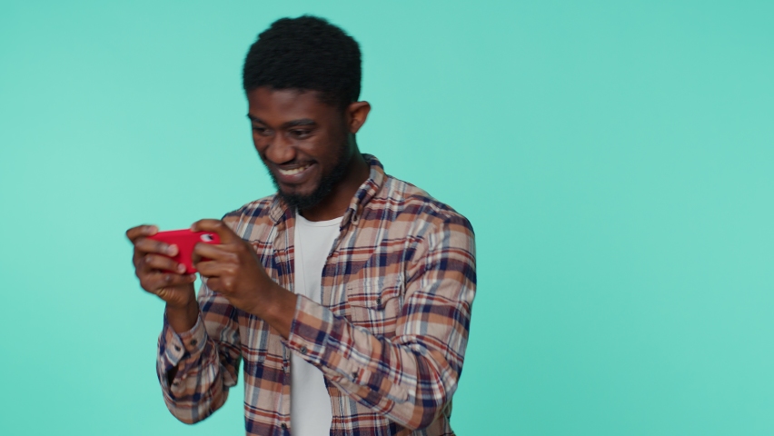 Worried funny addicted african american man enthusiastically playing racing video games on mobile phone. Young guy using smartphone gadget app with drive simulator isolayed on blue studio background