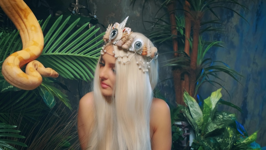 Close-up art portrait woman with snake. Nagga goddess touches white milk Boa constrictor. Fantasy asp girl blonde long hair, looks at serpent. Lady goth vampire queen viper. Backdrop green palants. 4k