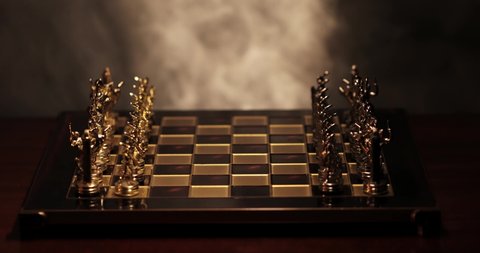 Chernivtsi, Ukraine - 04 04 2019:  a chessboard with golden pieces in the dark against a background of smoke in the initial position of the game. the man's hand moves the pawn and starts the game.