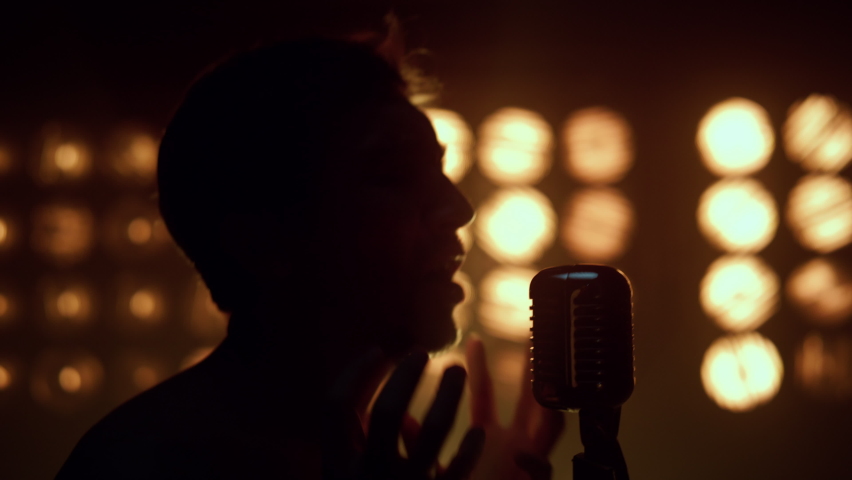 Unrecognizable man singing song microphone in night club spotlights. Close up silhouette of guy singer performing solo in studio lights. Professional vocalist making show on stage backlight indoors. Royalty-Free Stock Footage #1089197697