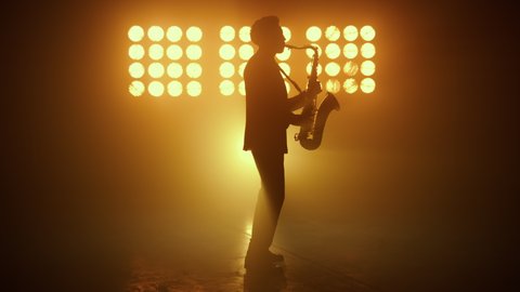 Silhouette of jazz musician playing saxophone in night club spotlights. Side view of unrecognizable stylish man performing melody on sax lit stage lights. Young guy saxophonist enjoying music indoors.
