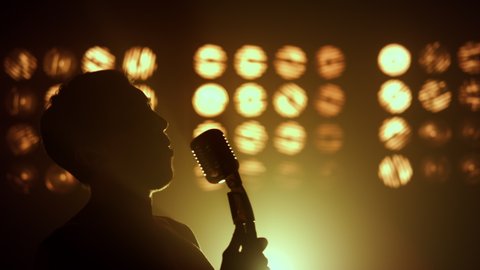 Professional singer performing song firely on stage nightclub close up. Silhouette unknown man performer raising mic on scene lights. Emotional guy vocalist singing microphone solo on show party. 