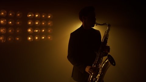 Silhouette of man saxophonist playing instrument on stage night club. Professional musician performing on golden saxophone on music concert indoor. Unknown stylish guy holding sax in studio spotlights