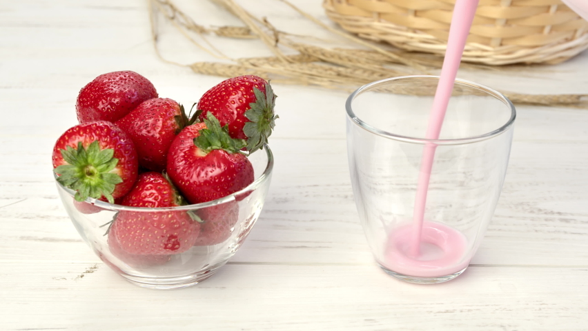 Pouring fresh creamy yogurt into glass with heap of seasonal ripe strawberries in bowl on wooden rustic table. Stream of dairy yoghurt vitamin appetizing summer breakfast brunch dessert delicious food
