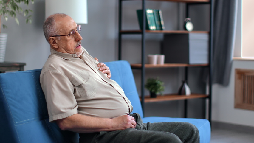 Senior man pressing hand chest heart attack suffering from unbearable pain sitting on couch at home. Elderly male having cardiac medical health problem painful sickness symptoms myocardial infarction | Shutterstock HD Video #1089199393