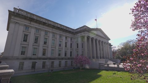 The north entrance of the U.S. Department of the Treasury Building in downtown Washington, D.C. seen on a sunny spring day. A tree with blossoming pink flowers is seen in the foreground.