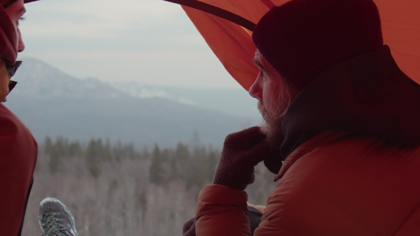Couple of tourists sitting together in tent on top of mountain and discussing scenic view while camping in winter