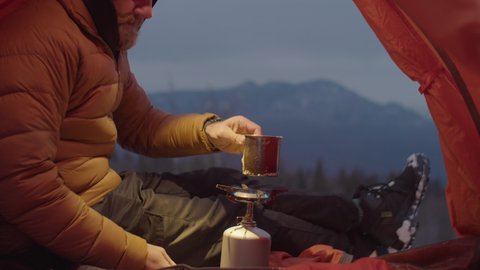 Male tourist preparing tea with portable gas stove and warming and by flame while sitting in camping tent on mountain top in winter: film stockowy