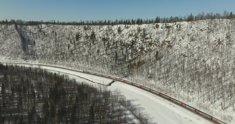 Passenger train an electric locomotive under the rock near river by two-sided winding Trans Siberian railway in the Ural Mountains. Aerial drone into the distance view at winter sunny day