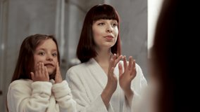 Content young woman and little daughter in white bathrobes applying cream on cheeks during skin care procedure and smiling with satisfaction while spending time together at home - Powered by Shutterstock - Get 15% off with code: PIKWIZARD15