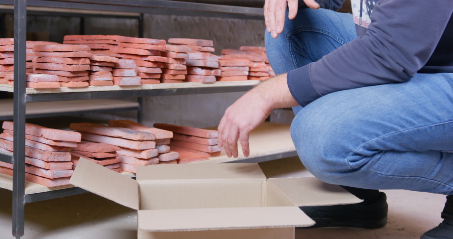 Person packs tile imitating old brick, shifting it from the shelf of rack into cardboard box to send delivery to customer or transport it to retail stores. Online trading in interior workshop