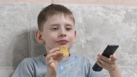 Cheerful caucasian boy 8 years old sitting on the couch eats potato chips, holds the TV remote control in his hand and switches the TV programs. The child watches TV. Schoolchildren's home rest