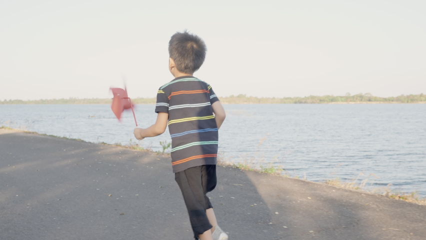 Asian child boy running and playing with pinwheels, Happiness little boy smiling in wheat field holding small wind wheel or windmill toy on hand in summer day at sunset in the park garden