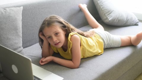 Cute child girl using laptop, lie on belly on grey couch, little smart kid typing on computer chatting with friend, talk online, study at home, playing and watch cartoons on gadget