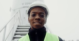 African-American man in a green vest and hard hat smiles with his teeth looking at camera, standing near the entrance to an industrial wind turbine, close-up, dynamic video. Video in 4k, red komodo