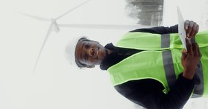 A young African-American man in work uniform with hard hat on his head smiles while looking at camera while standing near a wind turbine on a winter snowy day, vertical view. Video in 4k, red komodo