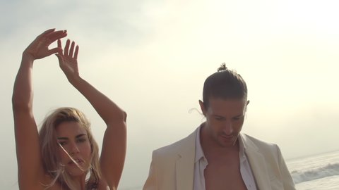 A couple of beautiful stylish lovers on the shore posing by the ocean on Malibu beach at sunset Slow motion.big waves A guy in a white suit looks at a girl and a girl in beautiful sexy clothes looks a