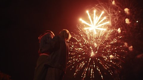 Young couple in love are standing outdoors against background of night sky and fireworks, back view. Couple of men and women stand against sky with fireworks