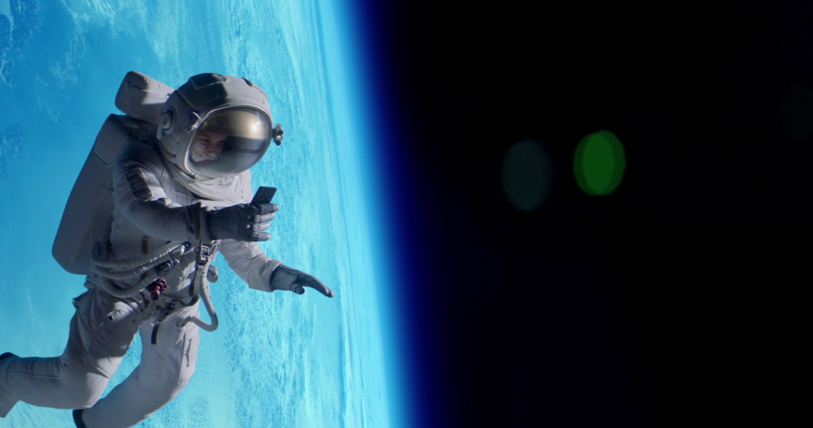 Female astronaut having a video call on her phone while performing space walk in open space, Earth in the background. Shot with 2x anamorphic lens Royalty-Free Stock Footage #1089214633