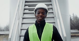 A young African-American man in a white hard hat and green vest looks at the camera with a smile, standing near the entrance to the wind turbine. Video in 4k, red komodo