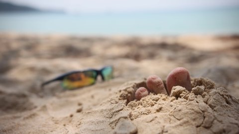 Tropical beautiful beach with sand, foot sticking out of the sand and sunny sunglasses. Summer travel or vacation concept.