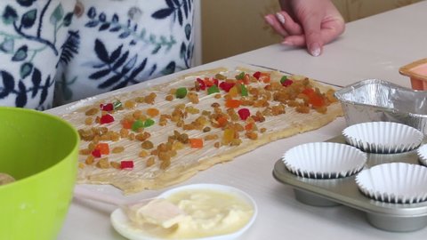 A woman pours raisins, candied fruits and almond flakes into a buttered rolled dough. Prepares cruffin with raisins and candied fruit. Tools and ingredients are laid out on the table.