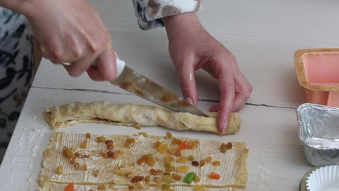 A woman cuts a roll of rolled dough. Prepares cruffin with raisins and candied fruit. Tools and ingredients are laid out on the table.