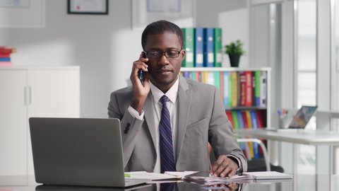 Portrait of African-American attorney advising client on smartphone and working on laptop. Entrepreneur having phone call with partner sitting at desk