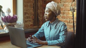 Beautiful African American woman in headwrap typing on laptop and then answering video call while working remotely in cafe