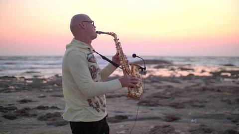 In summer, a young bald man saxophonist plays golden alt saxophone with a microphone, at sunset, on seashore, beautiful waves with white foam, against sea, in yellow sweater, black shorts. Cyprus.