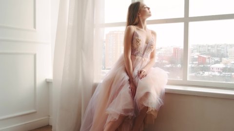 Portrait adult beautiful blonde girl princess sitting on windowsill. Young woman enjoys view. Large panoramic window white room silk curtain flutters waves in wind. Cute face, evening pink dress. 4k