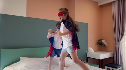 Two children in red and blue Superman costume,girl and boy superheroes, are jumping in room on bed,in children's room.Superheroes, brother and sister, play at home imagining they are superheroes.